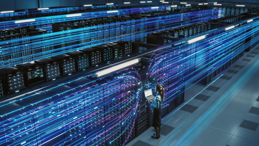 A woman in business attire stands in a futuristic data center filled with server racks, holding a laptop as glowing blue and purple digital lines representing data streams flow around her.