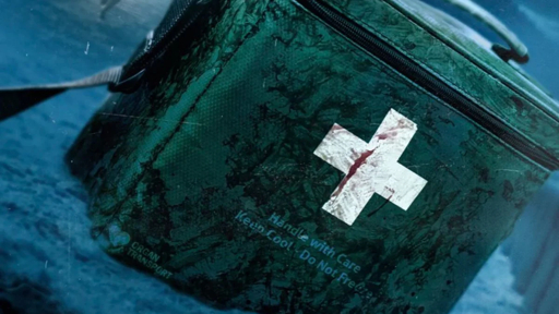 A close-up of a weathered green first aid bag with a white cross symbol stained with red, floating in blue water, featuring text “Handle with Care Keep Cool - Do Not Freeze” and a “COCON TRANSPORT” logo.
