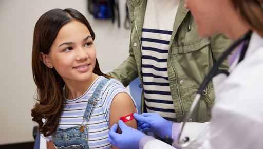A smiling young woman with long brown hair receives a vaccination in her upper arm from a doctor wearing blue gloves and a white coat, while another woman stands supportively behind her in a medical office setting.