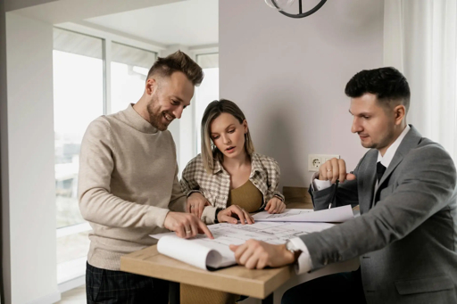 A male real estate agent in a suit consults with a young couple, pointing at architectural plans on a table in a modern home interior.