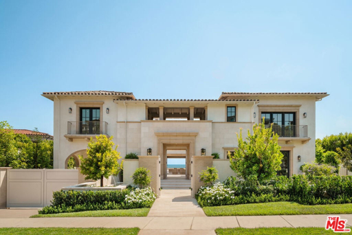 A large two-story beige stucco suburban home with a central arched entryway, balconies, manicured landscaping, and an ocean view in the background under a clear blue sky