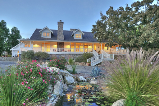 Luxury Santa Ynez ranch estate at dusk with stone chimney, wraparound porch, pond, lush gardens, and mature trees