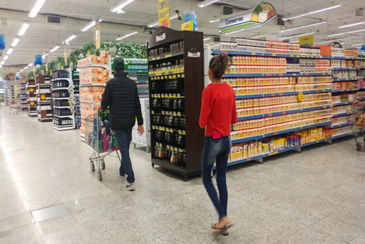 A man pushing a shopping cart and a woman walking in a brightly lit supermarket aisle stocked with products on shelves.