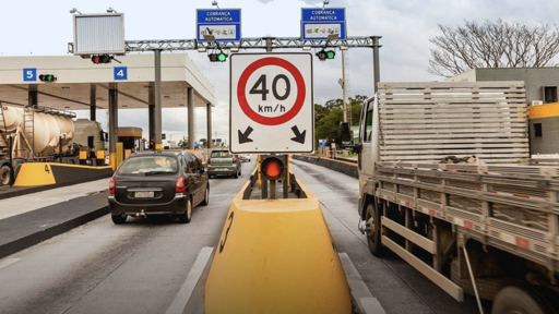 A Brazilian toll plaza on a highway with vehicles including a black car and a large truck approaching under a 40 km/h speed limit sign and overhead electronic signs.