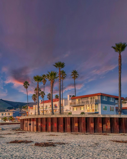 Row of multi-story beachfront homes with red roofs and balconies behind wooden seawall on Pismo Beach sandy shore