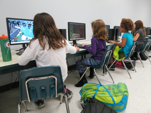 Four young girls use computers in an educational setting, with one playing Minecraft