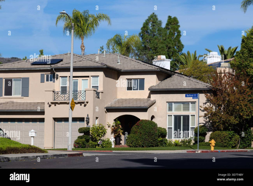A two-story beige stucco house with a gray shingled roof, solar panels, arched entryway, and attached garage, situated on a sunny suburban street lined with palm trees, bushes, and other homes under a clear blue sky.