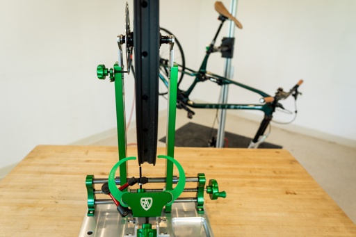 A bicycle wheel mounted in a precision truing stand on a workshop bench.