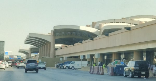 The image shows the exterior of King Khalid International Airport in Riyadh, featuring its distinctive curved beige terminal building with large arches, a multi-level roadway in the foreground with vehicles including cars, vans, and a black SUV, construction barriers, and workers in high-visibility vests.
