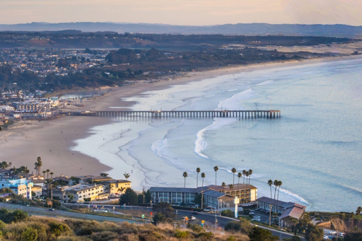 Aerial view of Pismo Beach Pier extending into the ocean, with sandy beach, waves, coastal buildings, and distant hills under a clear sky