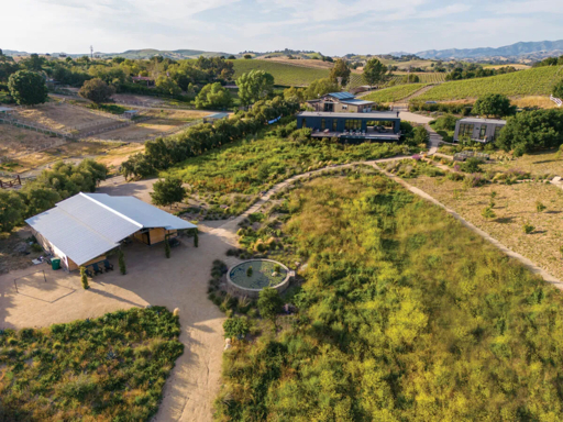 Aerial view of a modern Santa Ynez Valley ranch with house, metal-roofed barn, circular fountain, gardens, vineyards, and rolling hills
