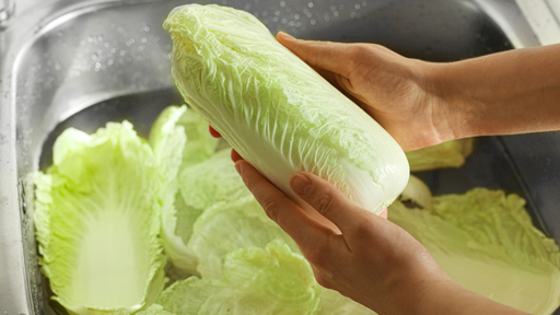A pair of hands holds a head of fresh Chinese cabbage under running water in a stainless steel sink, with loose leaves visible in the water below.