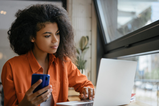 Femme professionnelle concentrée, utilisant un ordinateur portable et un smartphone dans un bureau moderne pour organiser sa recherche d'emploi avec l'IA