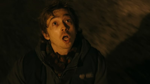 A close-up of a young man with messy brown hair, wide eyes, and an open mouth in a surprised expression, wearing a dark hooded jacket, set against a dark, blurry background with warm lighting.
