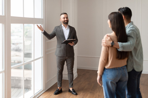 A male real estate agent in a suit holding a tablet and gesturing towards a window while showing a house to a couple of buyers standing nearby